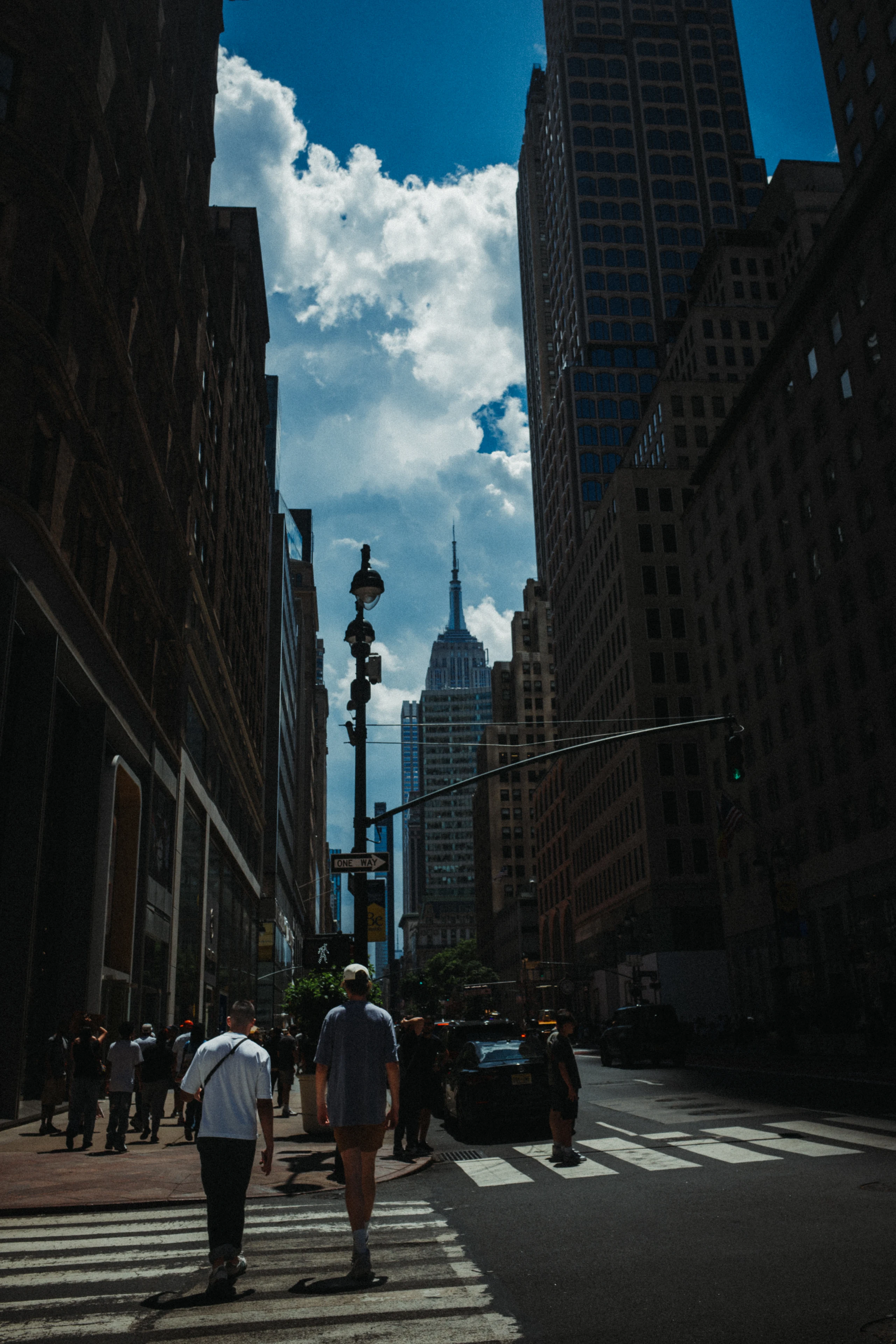 Pedestrians cross a shadowed Midtown street with the Empire State Building centered between tall buildings under blue sky and clouds.
