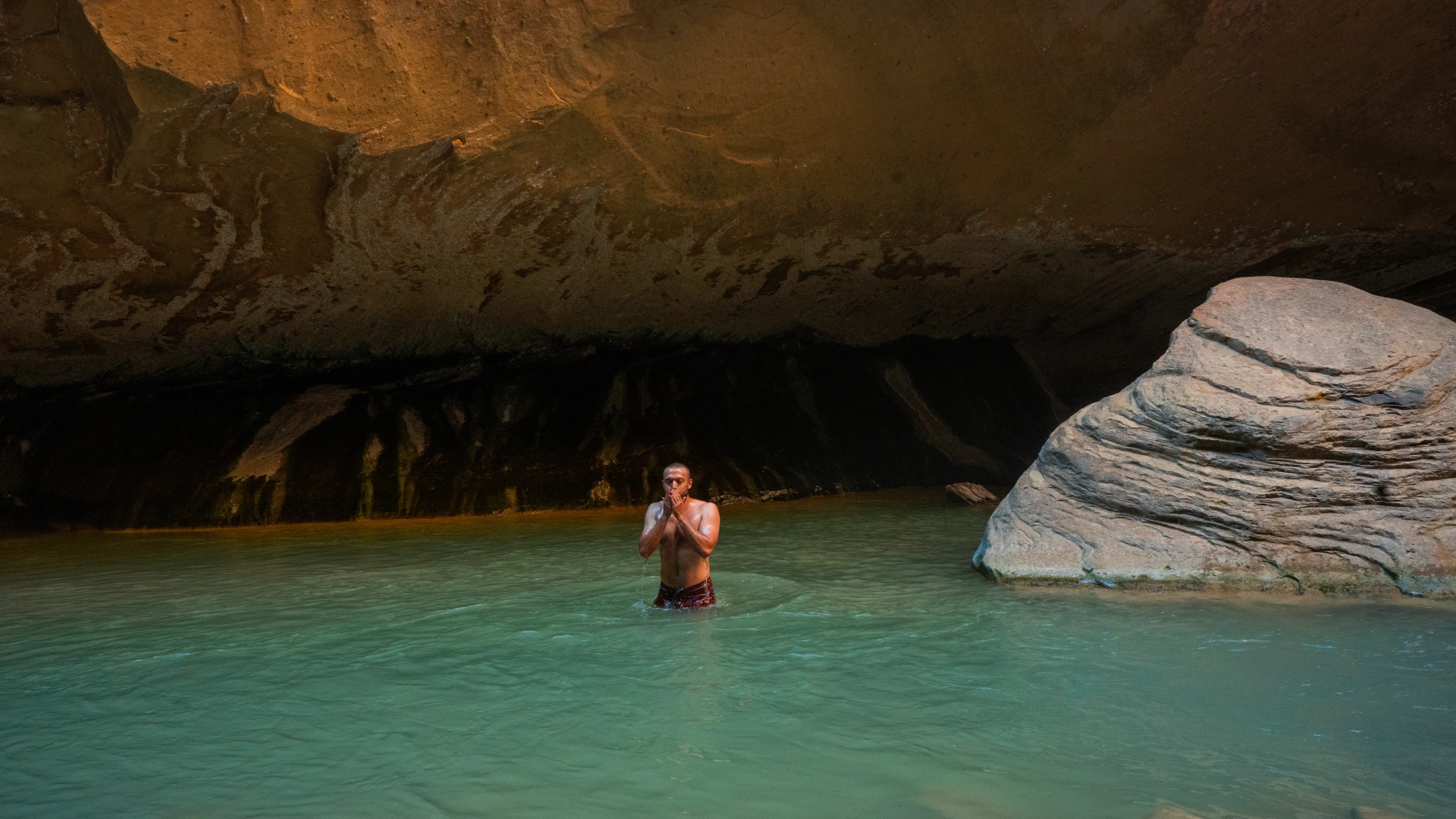 A person stands in a turquoise pool beneath a dark rock overhang in Zion.