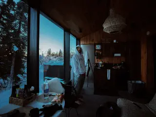 A person standing inside a dim cabin beside a wide window overlooking snowy trees.