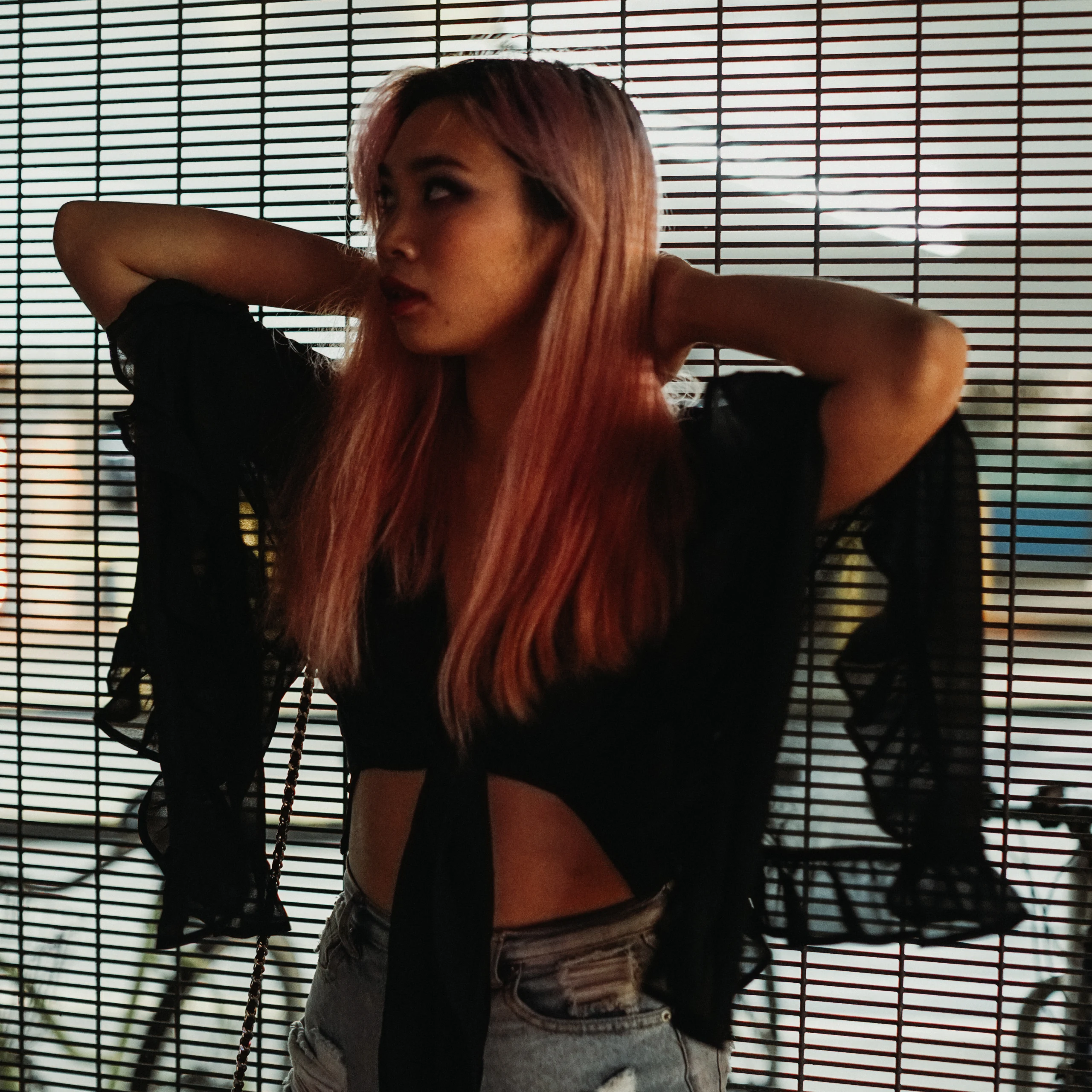 A woman with pink hair lifts her arms behind her head in front of a striped metal screen.