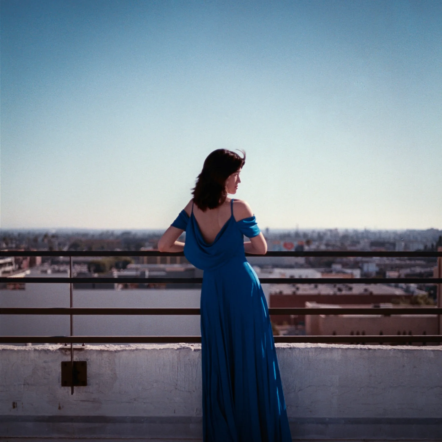 A woman in a long blue dress facing away at a rooftop railing.