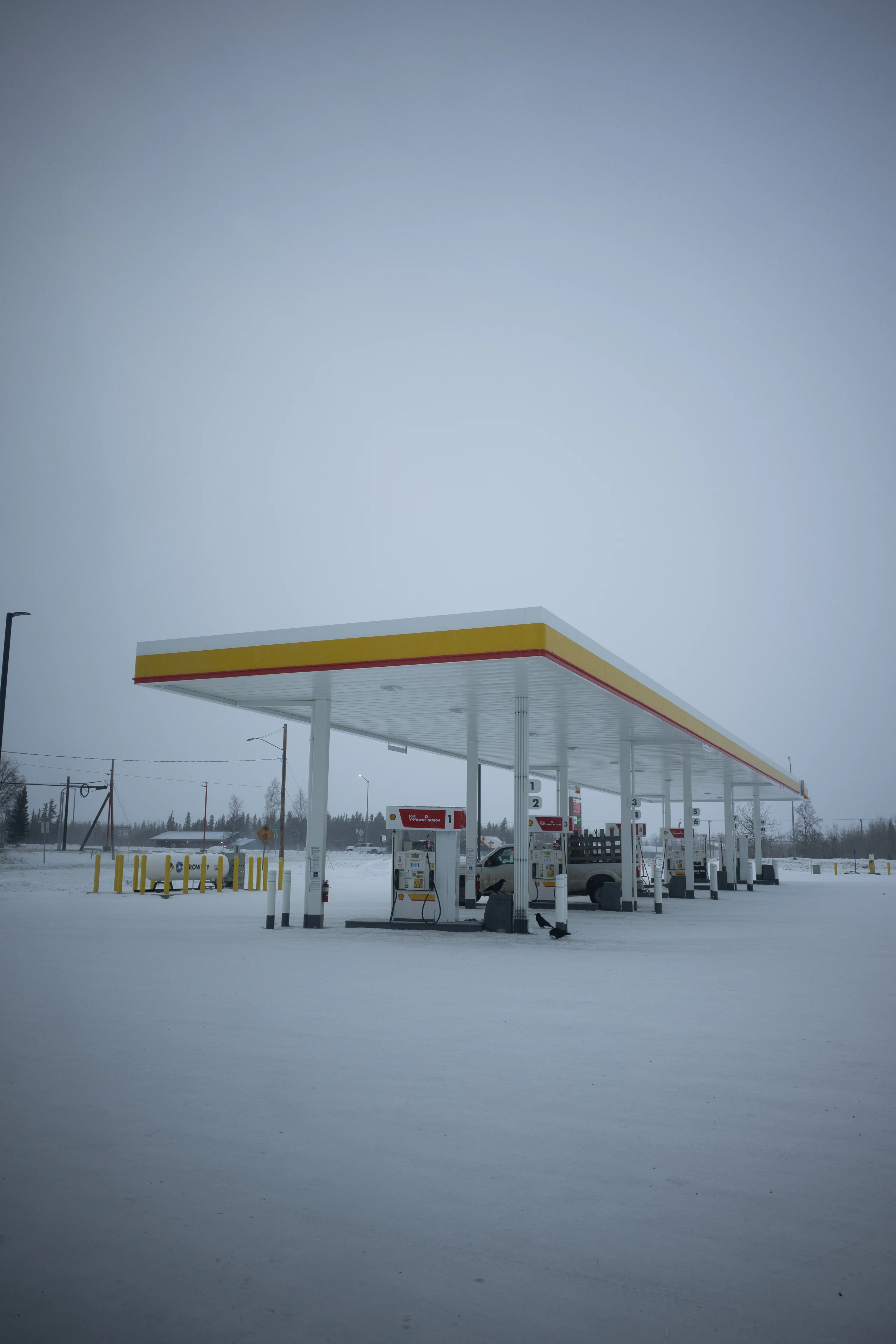 A yellow-and-red gas station canopy and fuel pumps stand in a snow-covered lot beneath a gray winter sky.