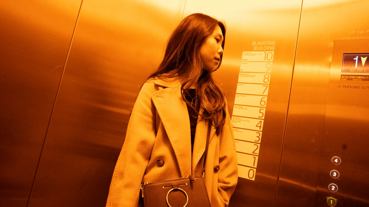 A woman in a camel coat stands inside a copper-colored elevator.