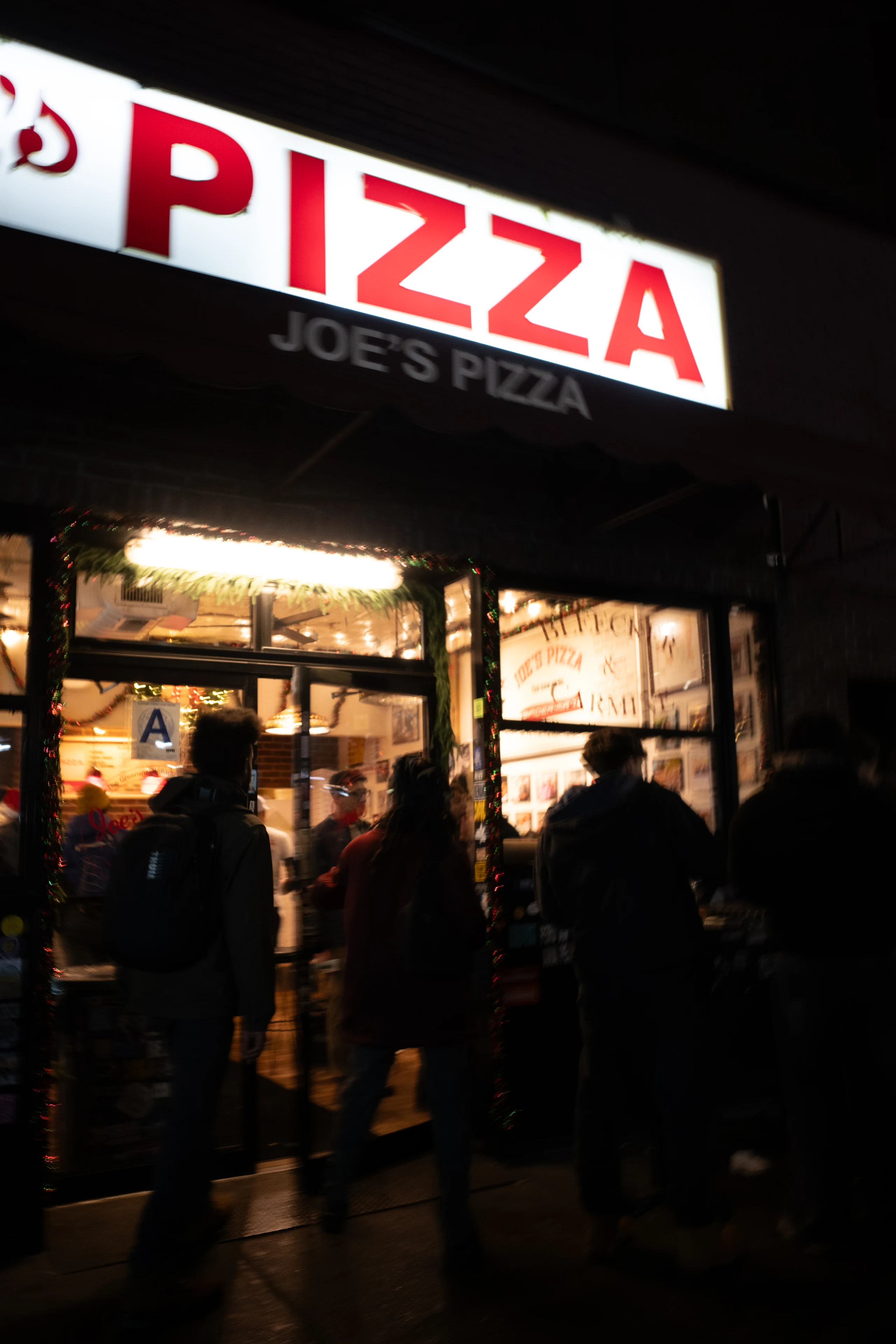 People stand outside Joe’s Pizza beneath its bright red-and-white sign at night.