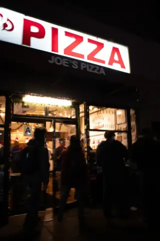 People stand outside Joe’s Pizza beneath its bright red-and-white sign at night.