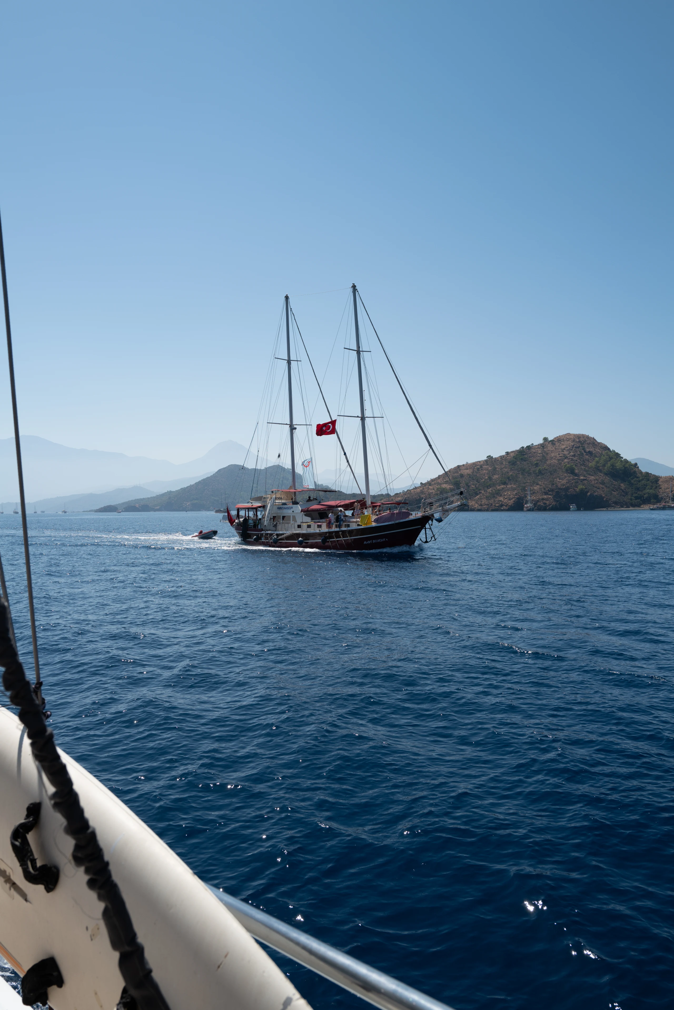 A traditional wooden sailing boat passes across bright water and layered hills.