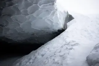 Dark, fractured glacier ice curves over a bright snowbank at the entrance to an ice cave.
