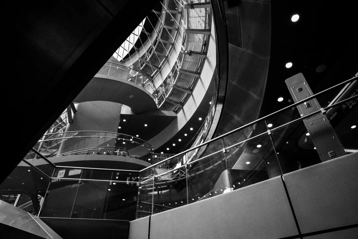 Black-and-white view looking upward through curving escalators, glass railings, structural beams, and ceiling lights.
