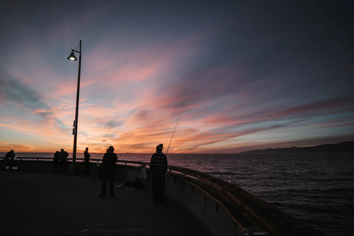 Silhouettes on a seaside promenade at sunset beneath a streetlamp.