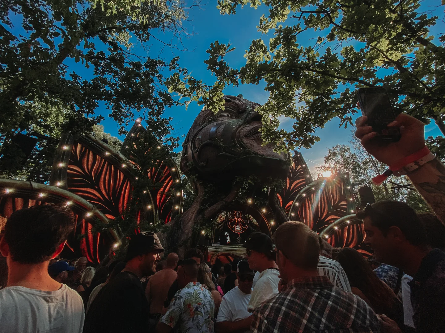 A crowd faces a large organic stage surrounded by leafy trees, red lights, and a bright blue afternoon sky.