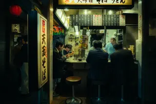Customers sit shoulder to shoulder at a brightly lit soba counter in Tokyo while a cook in a pale blue uniform looks across the small restaurant.