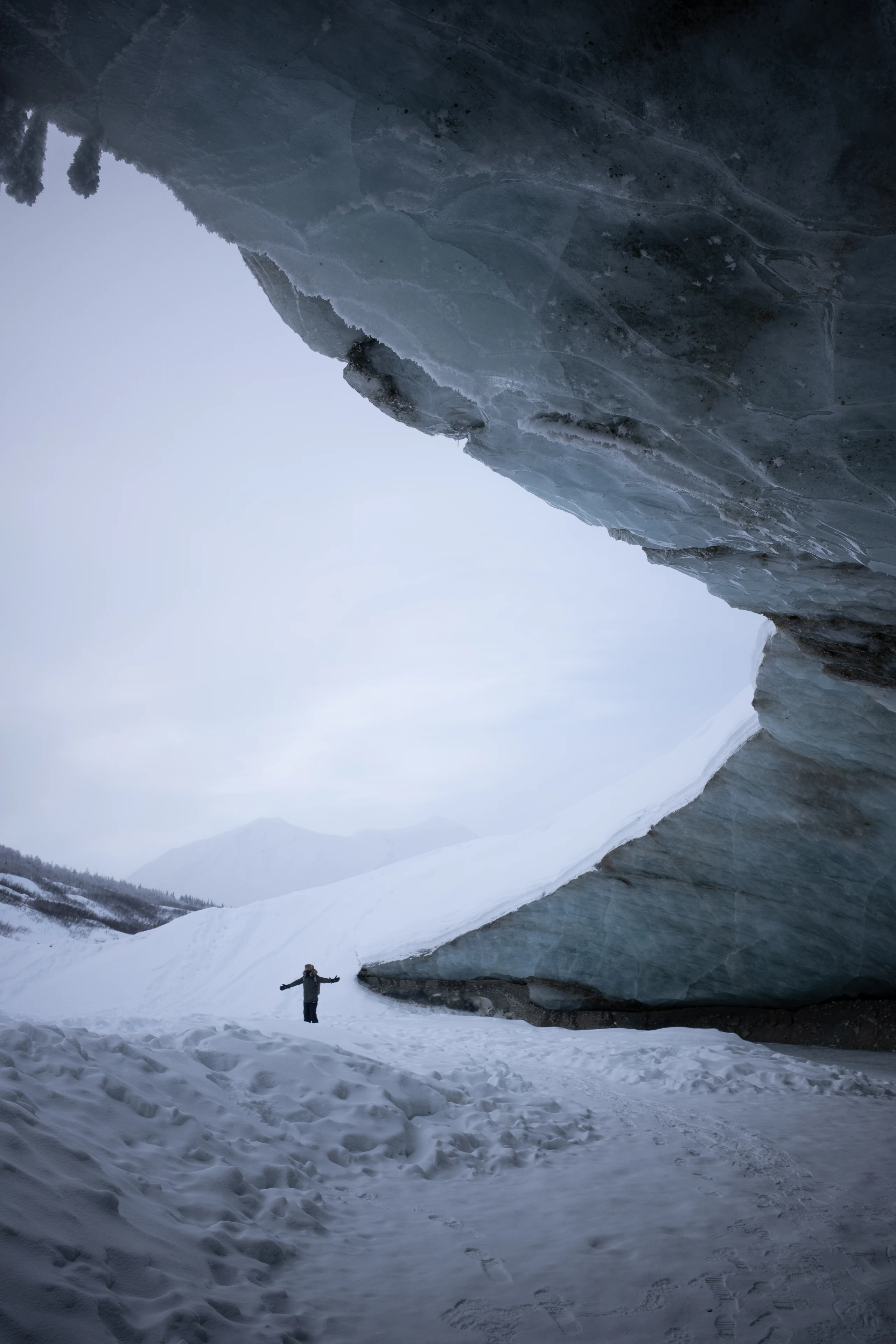 A person stands with arms outstretched below a dark glacier overhang framing snowy mountains.