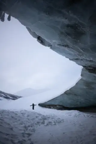 A person stands with arms outstretched below a dark glacier overhang framing snowy mountains.