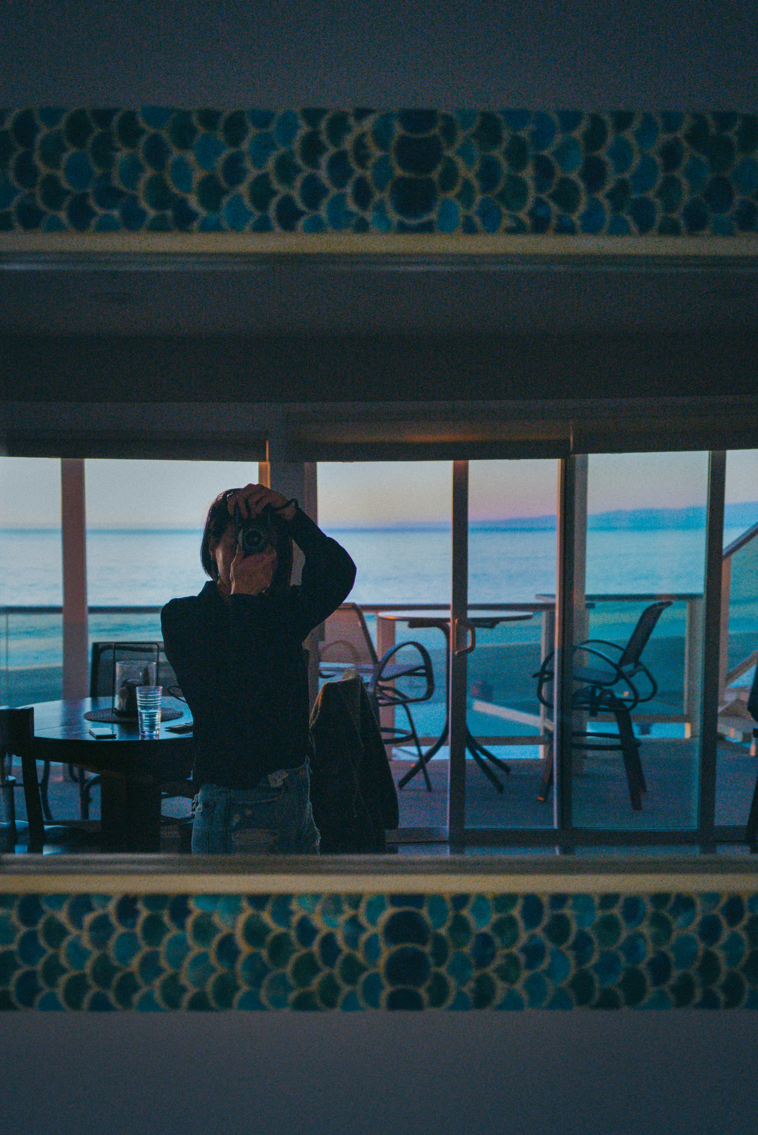 A silhouetted figure raises her hands to her hair in a tiled room overlooking the ocean.