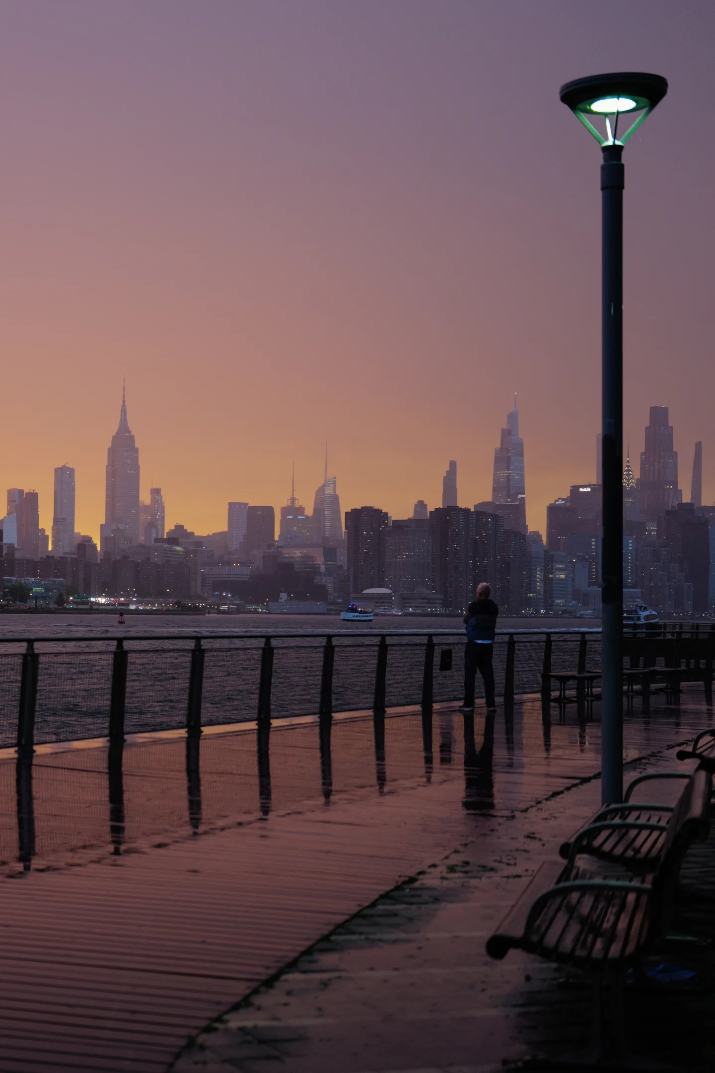 A lone figure on a rain-wet waterfront promenade facing the Manhattan skyline at dusk.