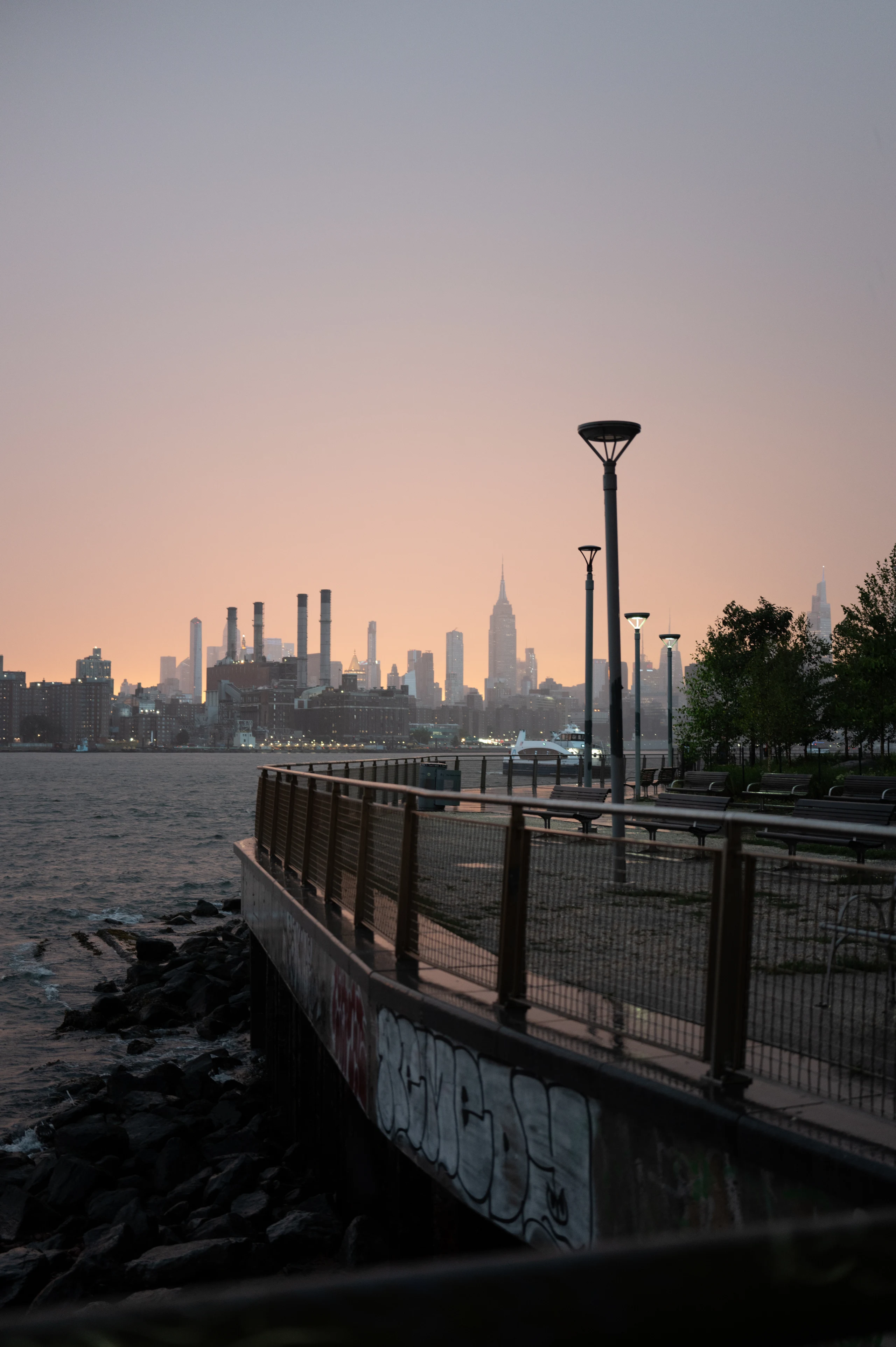 Waterfront path and lamps leading toward Manhattan beneath a pale pink evening sky.
